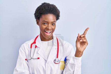 Foto de Mujer afroamericana vistiendo uniforme médico y estetoscopio sonriendo feliz señalando con la mano y el dedo hacia un lado - Imagen libre de derechos