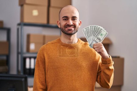 Photo for Young bald man with beard working at small business ecommerce holding money looking positive and happy standing and smiling with a confident smile showing teeth - Royalty Free Image