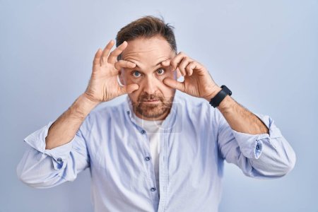 Photo for Middle age caucasian man standing over blue background trying to open eyes with fingers, sleepy and tired for morning fatigue - Royalty Free Image