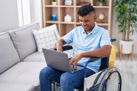 Téléchargez les photos : Jeune homme latin utilisant un ordinateur portable assis sur fauteuil roulant à la maison - en image libre de droit