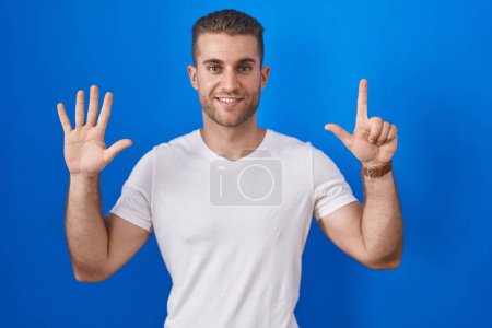 Foto de Joven hombre caucásico de pie sobre fondo azul mostrando y señalando hacia arriba con los dedos número siete mientras sonríe confiado y feliz. - Imagen libre de derechos