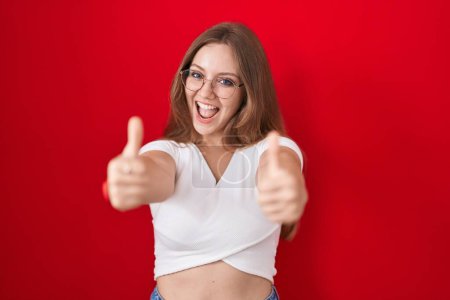 Téléchargez les photos : Jeune femme caucasienne debout sur fond rouge approuvant faire un geste positif avec la main, pouces levés souriant et heureux pour le succès. geste gagnant. - en image libre de droit