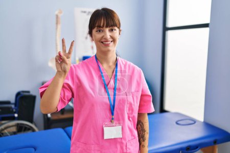 Foto de Joven morena que trabaja en la clínica de rehabilitación mostrando y señalando con los dedos número dos mientras sonríe confiada y feliz. - Imagen libre de derechos