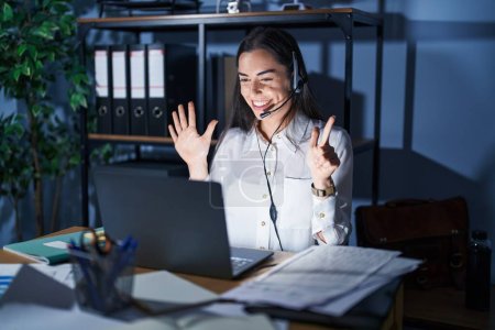 Photo for Young brunette woman wearing call center agent headset working late at night showing and pointing up with fingers number seven while smiling confident and happy. - Royalty Free Image