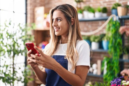 Téléchargez les photos : Jeune femme blonde fleuriste souriant confiant en utilisant un smartphone chez le fleuriste - en image libre de droit