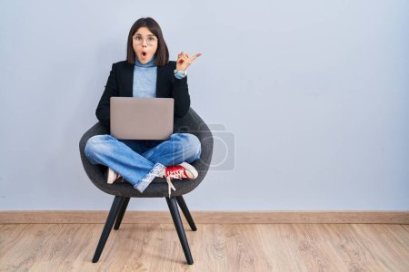 Foto de Mujer hispana joven sentada en silla usando computadora portátil sorprendida señalando con el dedo hacia un lado, boca abierta expresión asombrada. - Imagen libre de derechos