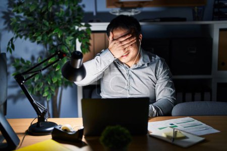 Téléchargez les photos : Jeune homme chinois travaillant à l'ordinateur portable la nuit souriant et riant avec la main sur le visage couvrant les yeux pour la surprise. concept aveugle. - en image libre de droit