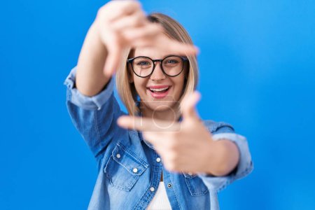 Téléchargez les photos : Jeune femme caucasienne debout sur fond bleu souriant faisant cadre avec les mains et les doigts avec le visage heureux. concept de créativité et photographie. - en image libre de droit