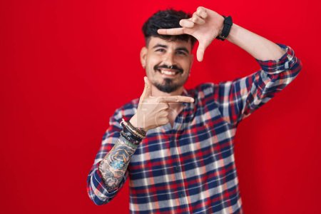 Téléchargez les photos : Jeune homme hispanique avec barbe debout sur fond rouge souriant faisant cadre avec les mains et les doigts avec le visage heureux. concept de créativité et photographie. - en image libre de droit