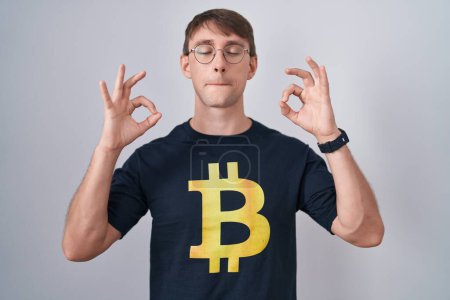 Téléchargez les photos : Homme blond caucasien portant Bitcoin t-shirt détendu et souriant avec les yeux fermés faire geste de méditation avec les doigts. concept de yoga. - en image libre de droit