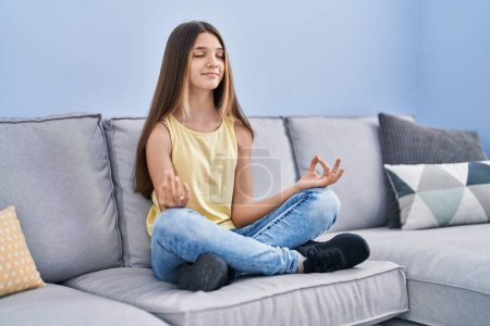 Foto de Adorable chica haciendo ejercicio de yoga sentado en el sofá en casa - Imagen libre de derechos