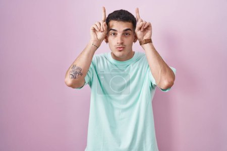Foto de Hombre hispano guapo de pie sobre fondo rosa haciendo gesto divertido con el dedo sobre la cabeza como cuernos de toro - Imagen libre de derechos