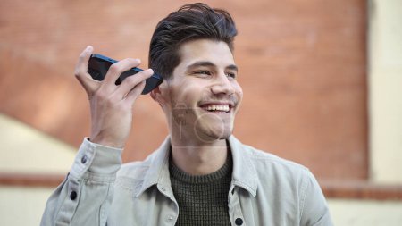 Foto de Joven hombre hispano sonriendo confiado escuchando mensaje de audio por el teléfono inteligente en la calle - Imagen libre de derechos