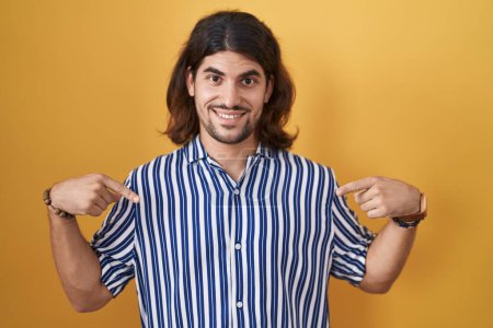 Foto de Hombre hispano con el pelo largo de pie sobre fondo amarillo mirando confiado con sonrisa en la cara, señalándose con los dedos orgullosos y felices. - Imagen libre de derechos