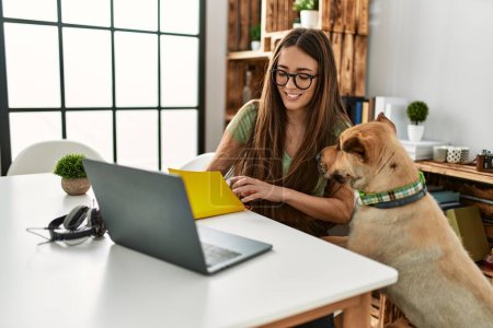 Photo for Young hispanic woman using laptop and writing on book sitting on table with dog at home - Royalty Free Image