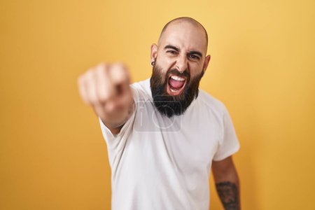 Photo for Young hispanic man with beard and tattoos standing over yellow background pointing displeased and frustrated to the camera, angry and furious with you - Royalty Free Image