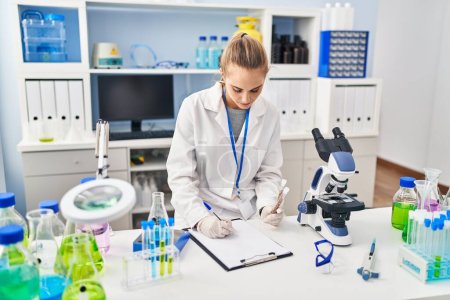Téléchargez les photos : Young blonde woman wearing scientist uniform holding test tubes writing on checklist at laboratory - en image libre de droit