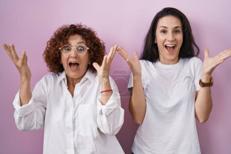 Téléchargez les photos : Hispanic mother and daughter wearing casual white t shirt over pink background celebrating crazy and amazed for success with arms raised and open eyes screaming excited. winner concept - en image libre de droit