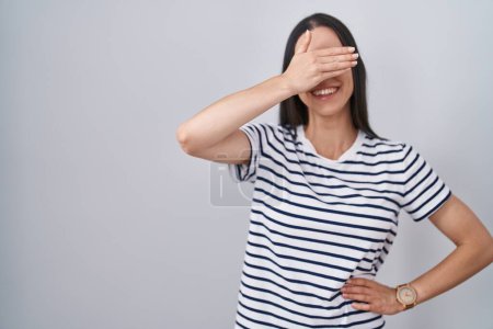Téléchargez les photos : Jeune femme brune portant un t-shirt rayé souriant et riant avec la main sur le visage couvrant les yeux pour la surprise. concept aveugle. - en image libre de droit