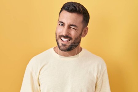 Téléchargez les photos : Bel homme hispanique debout sur fond jaune clin d'oeil en regardant la caméra avec expression sexy, gai et heureux visage. - en image libre de droit