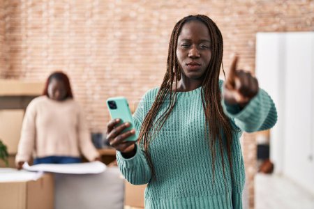 Téléchargez les photos : Femme africaine utilisant un smartphone à la nouvelle maison pointant du doigt la caméra et à vous, geste confiant regardant sérieux - en image libre de droit