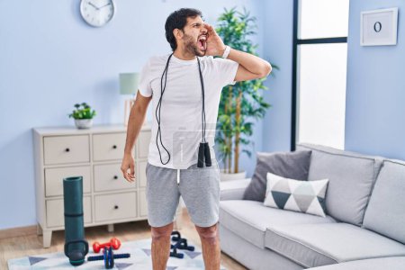 Foto de Hombre latino guapo usando ropa deportiva en casa gritando y gritando fuerte a un lado con la mano en la boca. concepto de comunicación. - Imagen libre de derechos