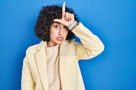 Photo for Young brunette woman with curly hair standing over blue background making fun of people with fingers on forehead doing loser gesture mocking and insulting. - Royalty Free Image
