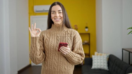 Photo for Young beautiful hispanic woman using smartphone doing ok gesture at home - Royalty Free Image