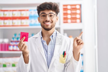 Photo for Arab man with beard working at pharmacy drugstore holding condom angry and mad screaming frustrated and furious, shouting with anger looking up. - Royalty Free Image