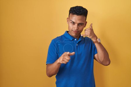 Photo for Young hispanic man standing over yellow background smiling doing talking on the telephone gesture and pointing to you. call me. - Royalty Free Image