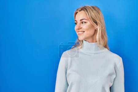 Foto de Young caucasian woman standing over blue background looking away to side with smile on face, natural expression. laughing confident. - Imagen libre de derechos