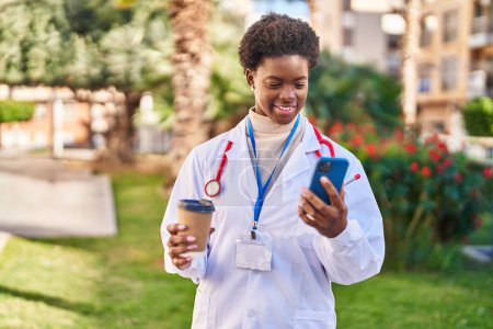 Téléchargez les photos : Femme afro-américaine portant l'uniforme de médecin en utilisant smartphone boire du café au parc - en image libre de droit