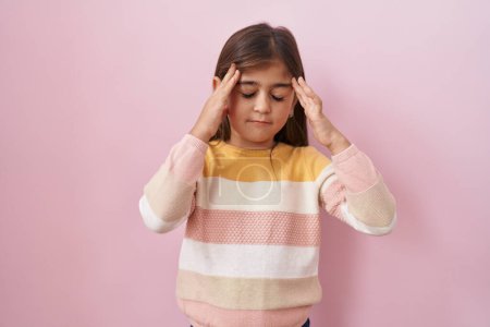 Téléchargez les photos : Petite fille hispanique portant un pull sur fond rose avec la main sur la tête, mal de tête parce que le stress. migraine souffrant. - en image libre de droit