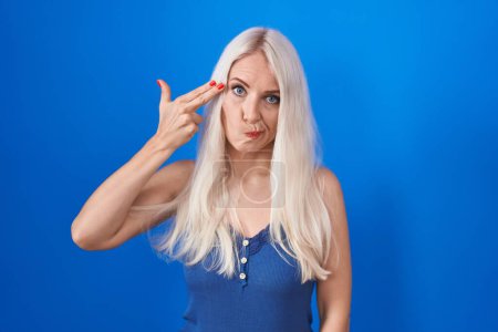 Téléchargez les photos : Femme caucasienne debout sur fond bleu tirant et se suicidant pointant la main et les doigts vers la tête comme une arme à feu, geste de suicide. - en image libre de droit
