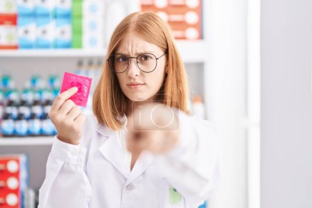 Téléchargez les photos : Jeune rousse femme travaillant à la pharmacie pharmacie tenant préservatif pointant du doigt la caméra et à vous, geste confiant regardant sérieux - en image libre de droit
