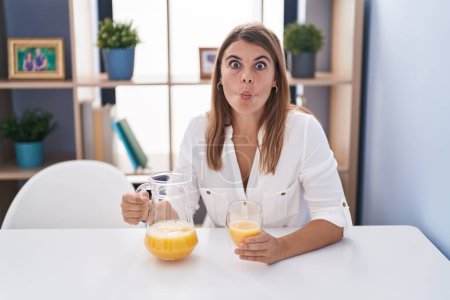 Téléchargez les photos : Jeune femme hispanique buvant un verre de jus d'orange faisant visage de poisson avec la bouche et les yeux plissés, fou et comique. - en image libre de droit