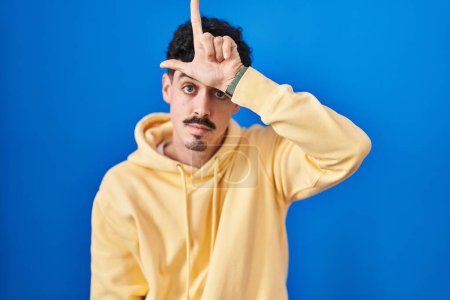 Photo for Hispanic man standing over blue background making fun of people with fingers on forehead doing loser gesture mocking and insulting. - Royalty Free Image