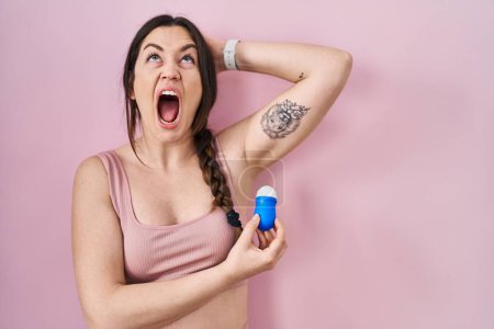 Foto de Mujer morena joven usando rollo en desodorante enojado y loco gritando frustrado y furioso, gritando con ira mirando hacia arriba. - Imagen libre de derechos