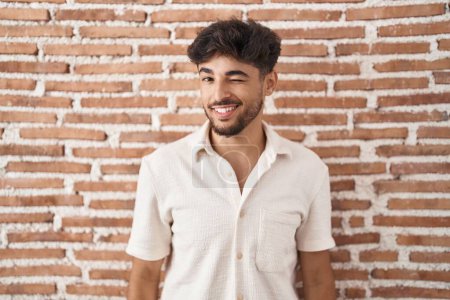 Téléchargez les photos : Arabe homme avec barbe debout sur le mur de briques arrière-plan clin d'oeil en regardant la caméra avec expression sexy, gai et heureux visage. - en image libre de droit