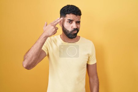 Téléchargez les photos : Homme hispanique avec la barbe debout sur fond jaune tirant et se suicidant pointant la main et les doigts vers la tête comme une arme à feu, geste suicidaire. - en image libre de droit