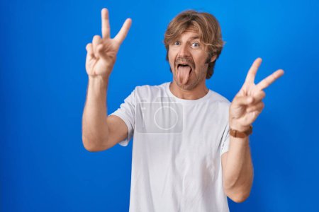 Foto de Hombre de mediana edad de pie sobre fondo azul sonriendo con la lengua hacia fuera mostrando los dedos de ambas manos haciendo signo de victoria. número dos. - Imagen libre de derechos