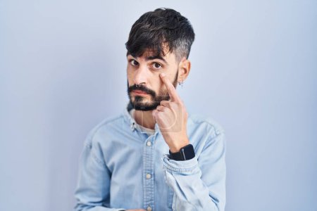 Téléchargez les photos : Jeune homme hispanique avec la barbe debout sur fond bleu pointant vers l'œil vous regardant geste, expression suspecte - en image libre de droit