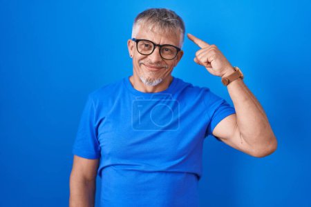 Téléchargez les photos : Homme hispanique avec les cheveux gris debout sur fond bleu souriant pointant vers la tête avec un doigt, grande idée ou pensée, bonne mémoire - en image libre de droit