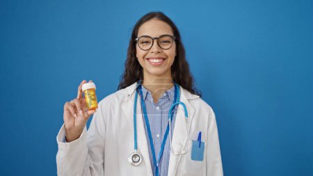 Téléchargez les photos : Jeune belle hispanique femme médecin tenant des pilules sur fond bleu isolé - en image libre de droit