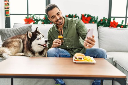 Téléchargez les photos : Jeune homme hispanique faire selfie assis sur le canapé avec chien par décor de Noël à la maison - en image libre de droit