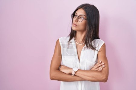 Téléchargez les photos : Brunette jeune femme debout sur fond rose portant des lunettes regardant sur le côté avec les bras croisés convaincu et confiant - en image libre de droit