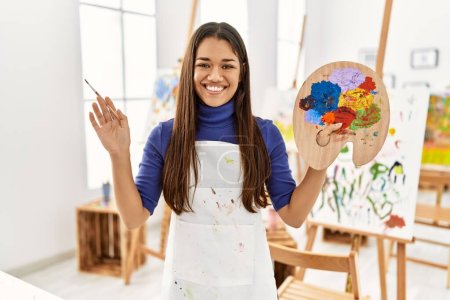 Foto de Mujer latina joven sonriendo confiada sosteniendo el pincel y la paleta en el estudio de arte - Imagen libre de derechos