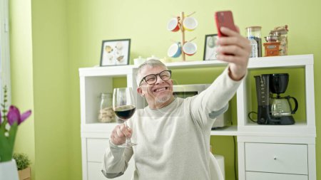 Foto de Hombre de pelo gris de mediana edad bebiendo copa de vino tomando selfie por teléfono inteligente en casa - Imagen libre de derechos