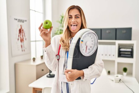 Photo for Young blonde doctor woman holding weighing machine and green apple sticking tongue out happy with funny expression. - Royalty Free Image