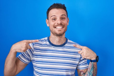 Photo for Young hispanic man standing over blue background looking confident with smile on face, pointing oneself with fingers proud and happy. - Royalty Free Image
